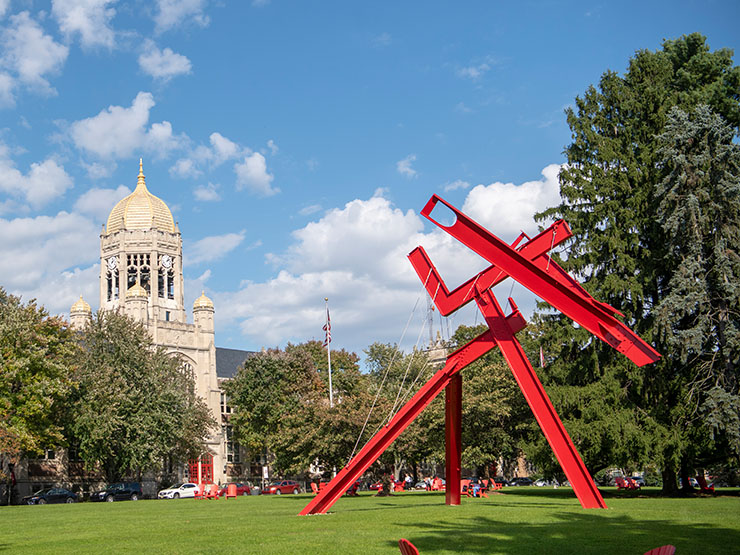 Red sculpture placed in foreground of college green with clocktower off into the background.