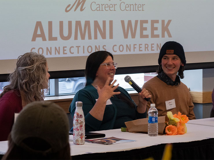 A group of adult professionals sit behind a long table and speak as a panel during an alumni networking event.