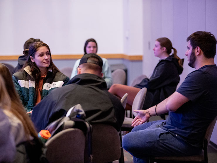 A group of young adults sit in chairs and talk with one another in a large, open room.