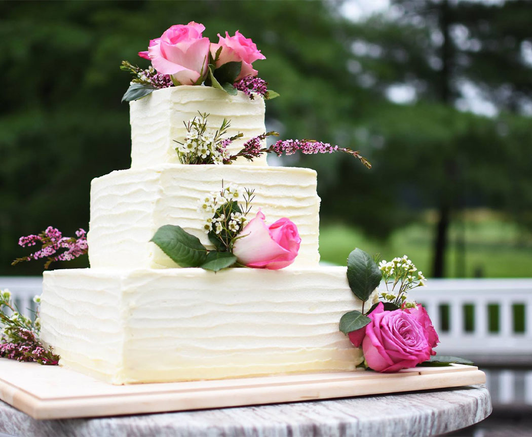 A three-tiered white wedding cake, adorned with pink roses, stands outdoors.