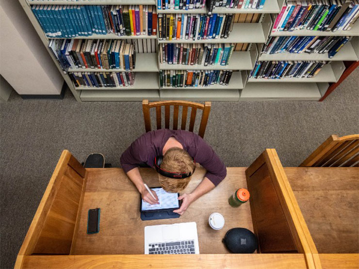 Photographed from above, a college student wearing headphones is seated a desk in a library is immersed in work.