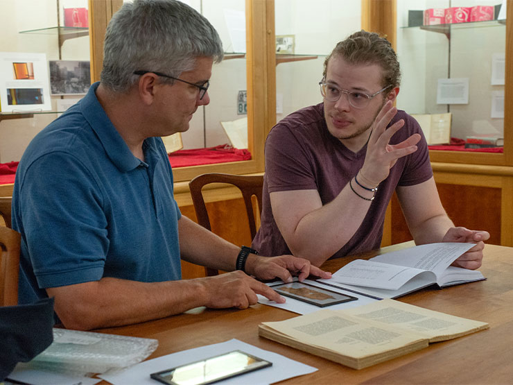 An adult and young adult hold a conversation at a table scattered with open books in an academic building.