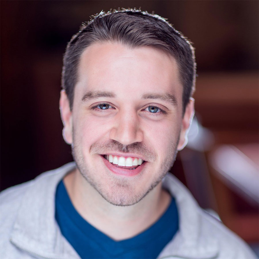 An adult in a blue shirt, with a grey shirt on top, smiles at the camera.