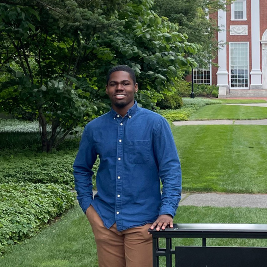 A student in a blue dress shirt and dark tan pants stands near a sign on a college green.