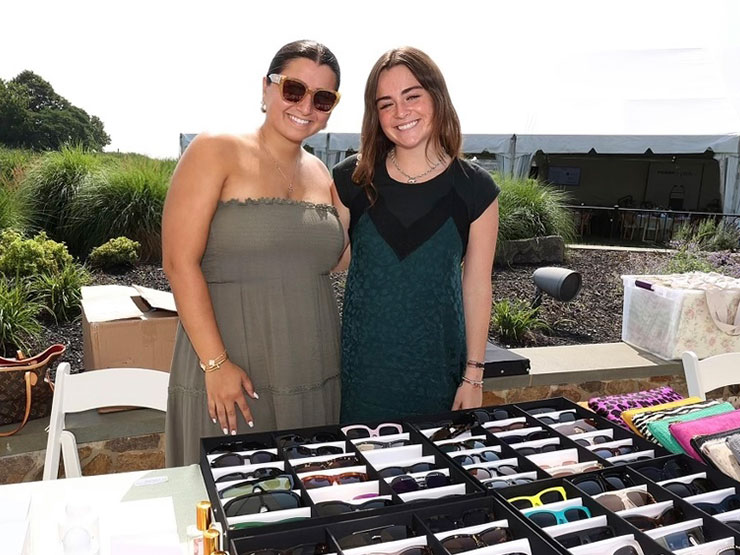 An adult woman and a young woman, both wearing dresses, stand behind a table containing accessories.