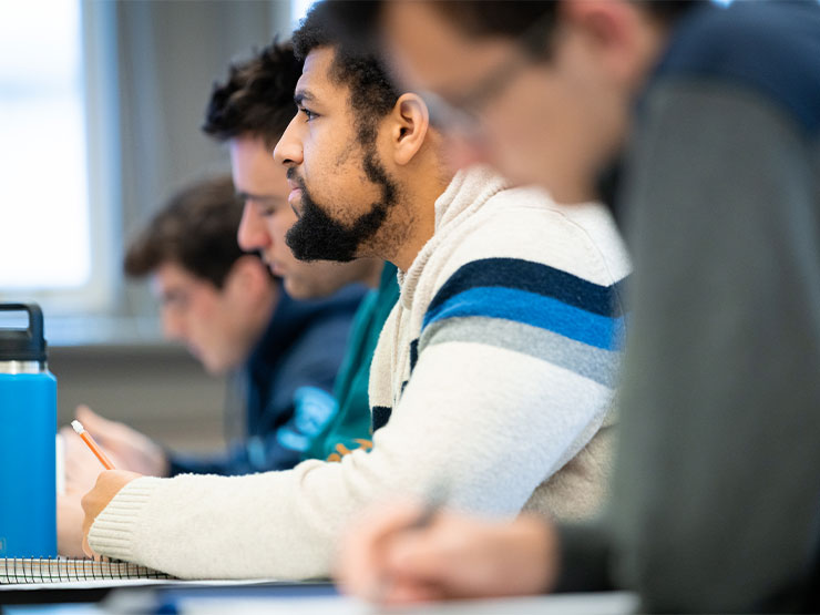 A row of young adults are seated at desks and look to the front of a classroom.