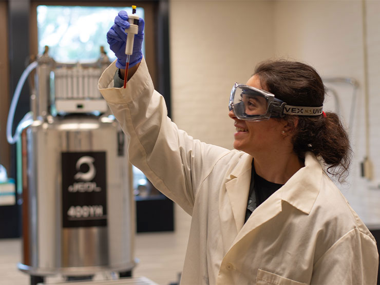 A college student in a white lab coat and goggles holds up a test tube in a laboratory.