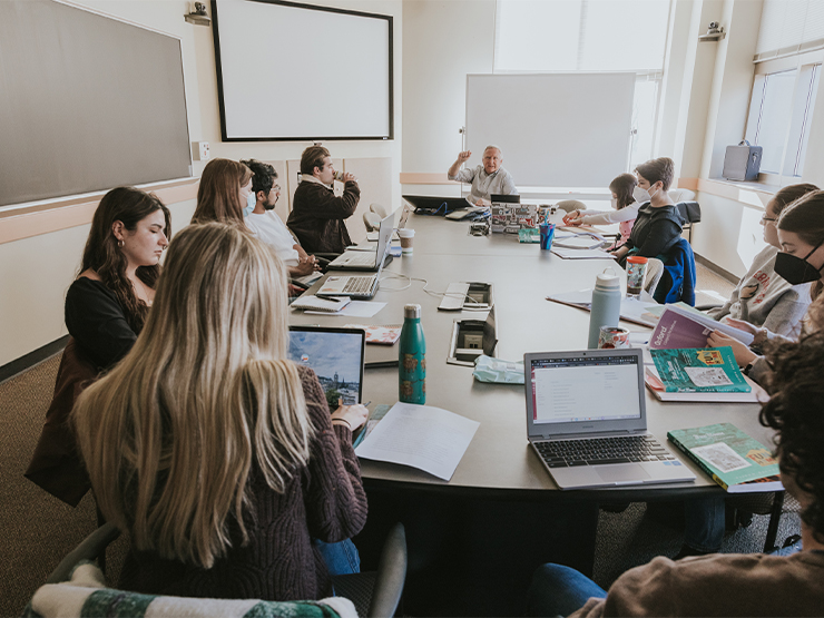 Students are gathered around a table, crowded with laptops, books and water bottles, in a classroom. A man sits at the end of the table and speaks to the group.