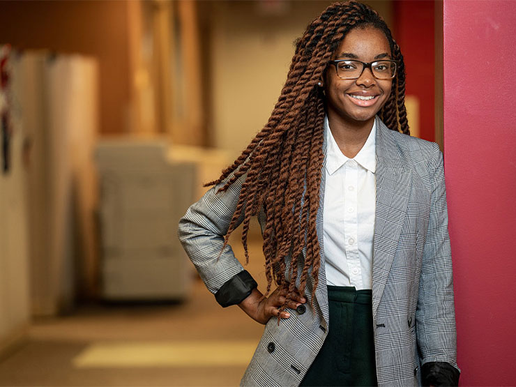 A young adult with long braided hair wears in a gray, black and white business suit and stands, hand on hip in the door way of a professional building.