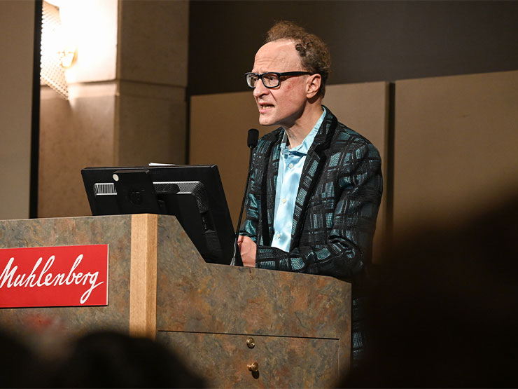 An adult in a plaid suit jacket speaks at a podium that has a placard that reads 