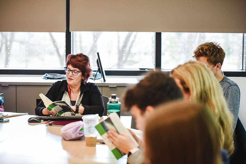 Faculty member lecturing in bright sunlit filled classroom amongst group of students seated at conference table.