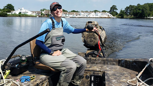 A young woman in chest waders sits on the back of a boat speed boat moving through a body of water.