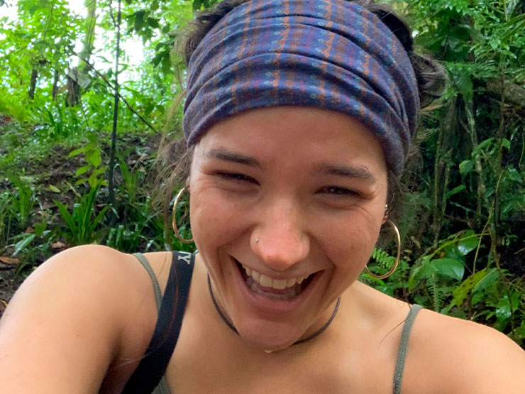 A young adult laughs and smiles at the camera while outdoors in a tropical environment.