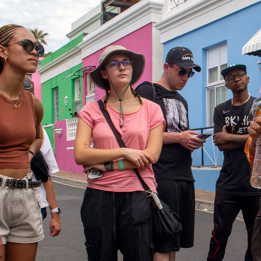 A young adult in a pink shirt and bucket hat looks to the distance while standing in a neighborhood full of colorful homes and other young adults nearby.