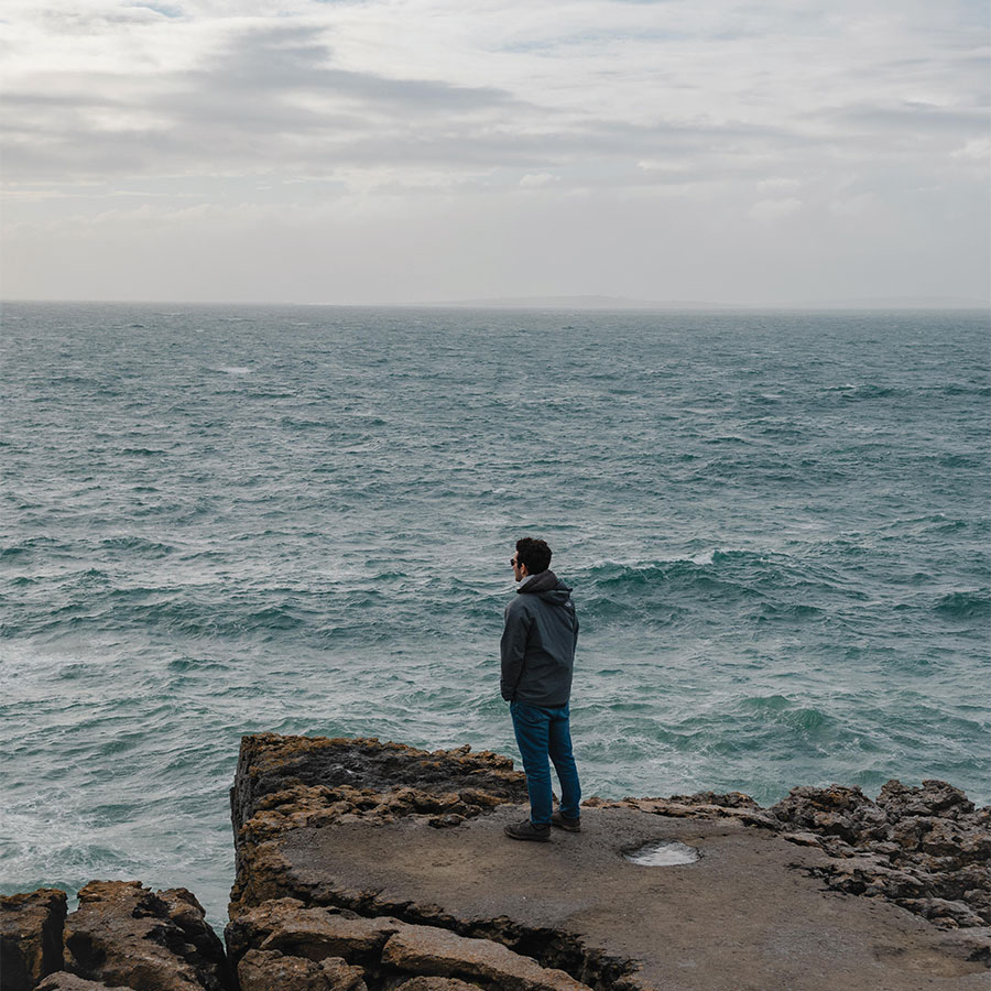 A young adult stands on a rock outcropping, staring out at the choppy waves of a blue-grey sea.