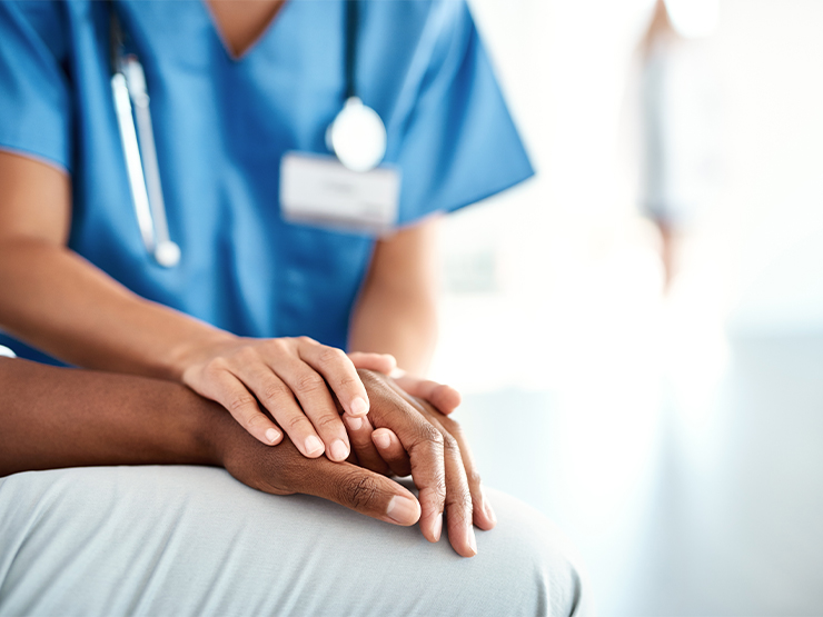 A person wearing blue hospital scrubs and a stethoscope lays their hands on the arm of another person in a comforting manner.