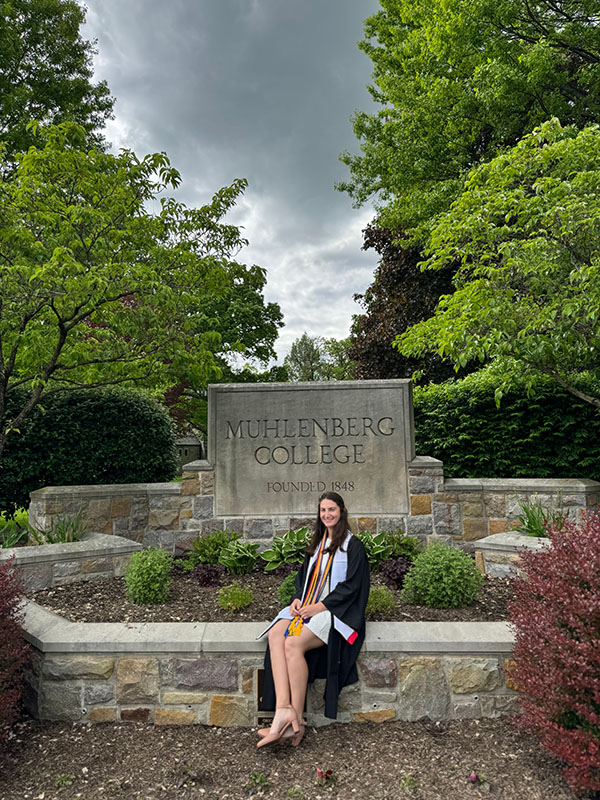 Muhlenberg History program alumni, Sophia Framm '24, seated outdoors on stone campus signage reading 