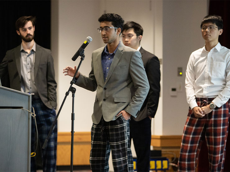 A group of college students wearing suits and plaid dress pants stand together at the front of a room, one of them speaking into a microphone.