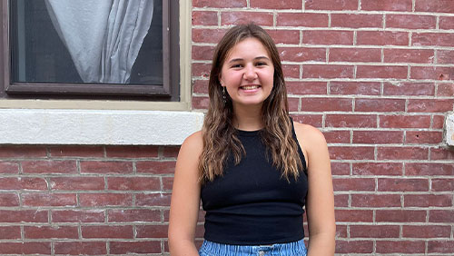 A young adult with long brown hair wears a black tank top and smiles for the camera against a red brick building.