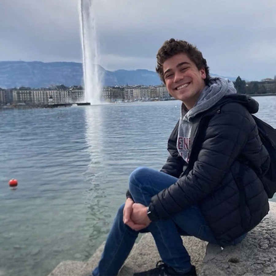 A young adult poses while seated on the ledge of a body of water with a large fountain in the distance.