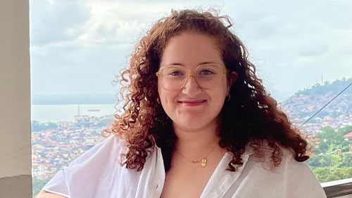 A woman with glasses and curly shoulder-length hair poses for the camera with the view of city sprawled out in the distance behind.