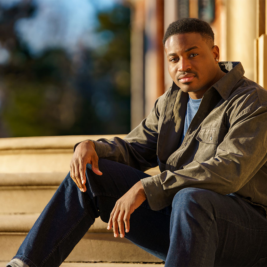 A man sits on stone steps in fading daylight, his hands resting on his knees while looking at the camera.