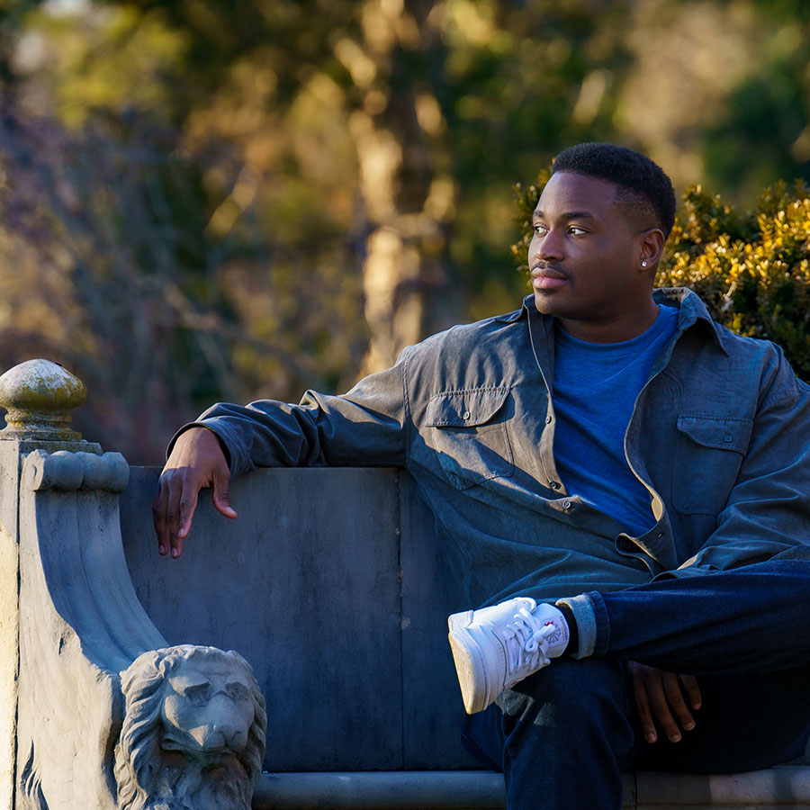 A man sits on casually on a stone bench in the fading daylight and looks off into the distance.