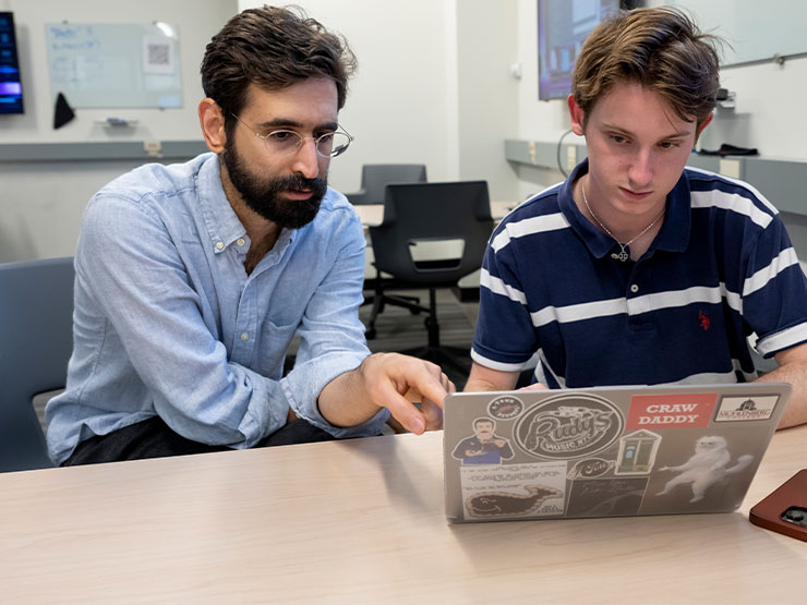 An instructor leans over the shoulder of a student with an open laptop, pointing at the screen.