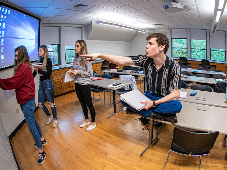 A group of students in a classroom look at a blackboard, while one of them, sitting on a desk points toward the board.