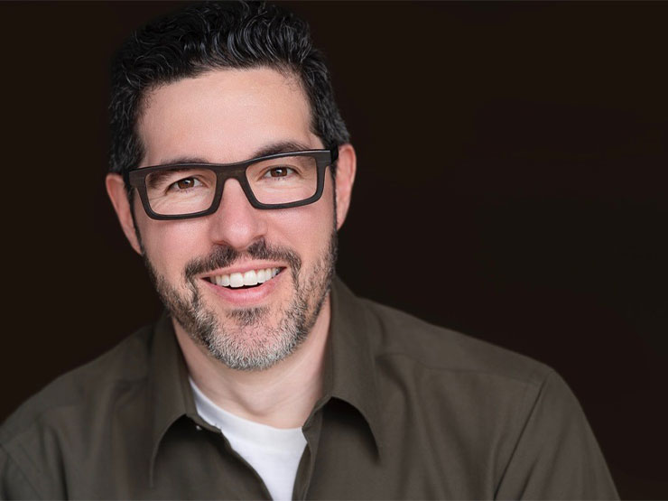 A professional headshot of an adult with glasses, wearing a brown dress shirt against a black background