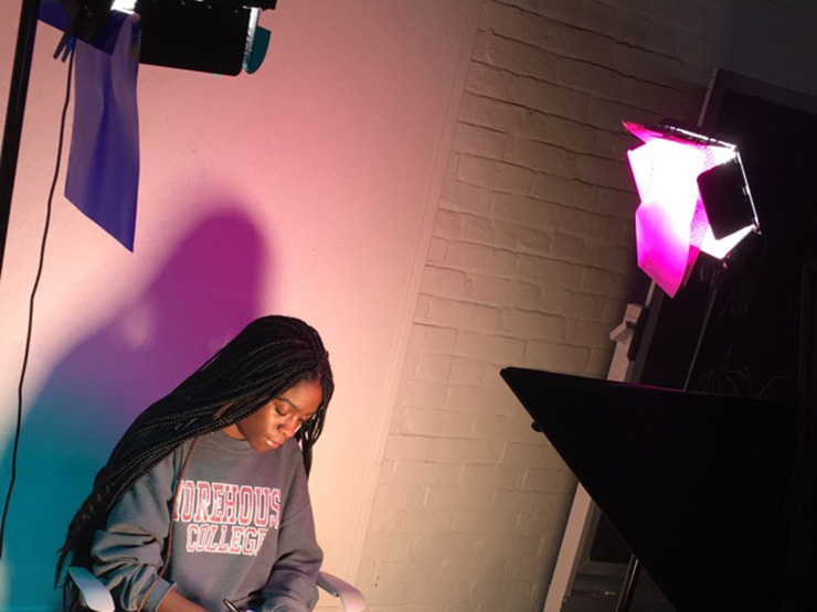 A college student sits against a wall lit with colorful lighting.