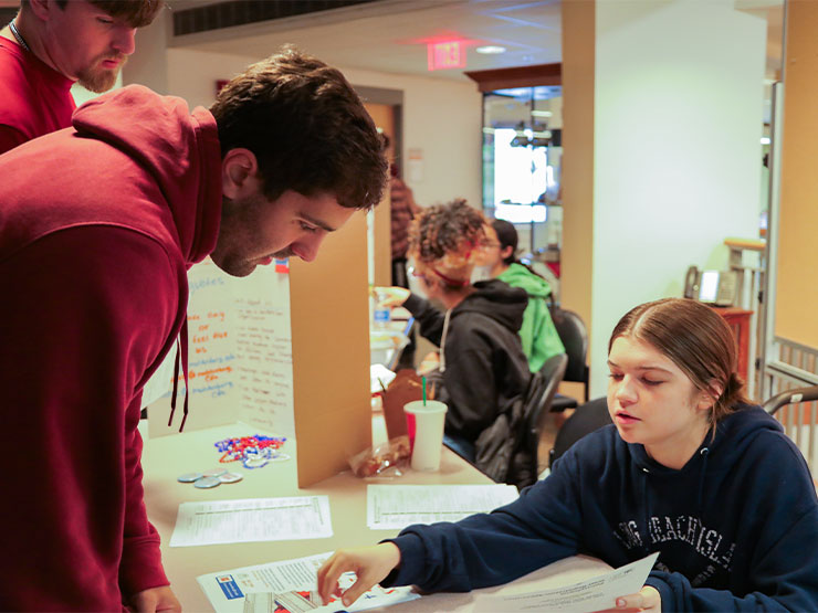 A college student seated at a table helps point out information on a sheet of paper to another student stopping by.