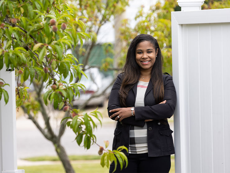 A woman in a black pantsuit stands outside, leaning on a white fence, and smiles at the camera