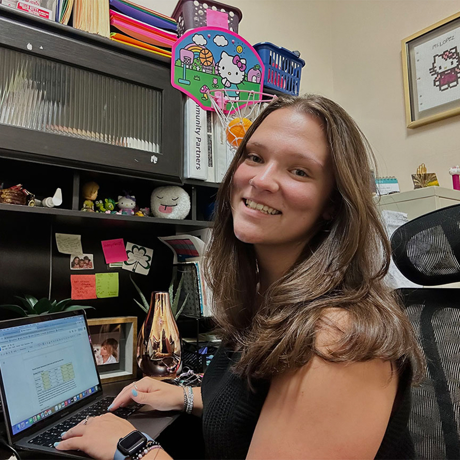 A young adult with long wavy brown hair smiles at the camera from a desk with an open laptop.