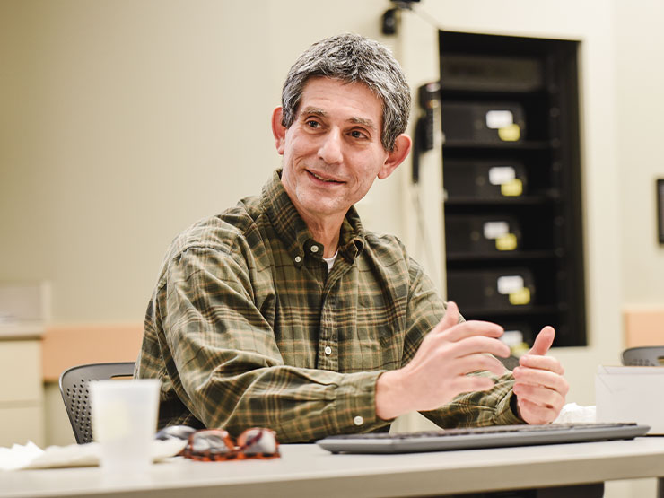 A college professor, seated at a table, in a smiles while speaking in a classroom.