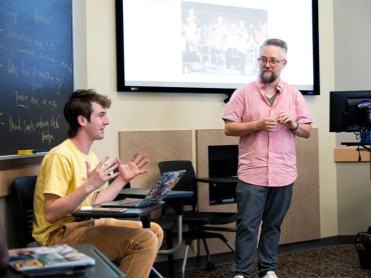 An instructor looks on as a seated students speaks in a classroom.