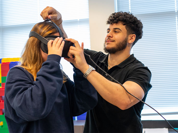 A college student helps a peer adjust a headset of virtual reality goggles in a learning environment.