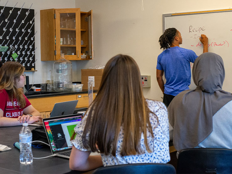 A group of students sit around a table in a lab as one of the students writes notes on a whiteboard.