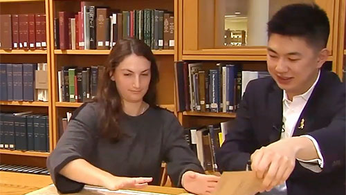 A pair of college students sit at a desk in a library and look through aging documents.