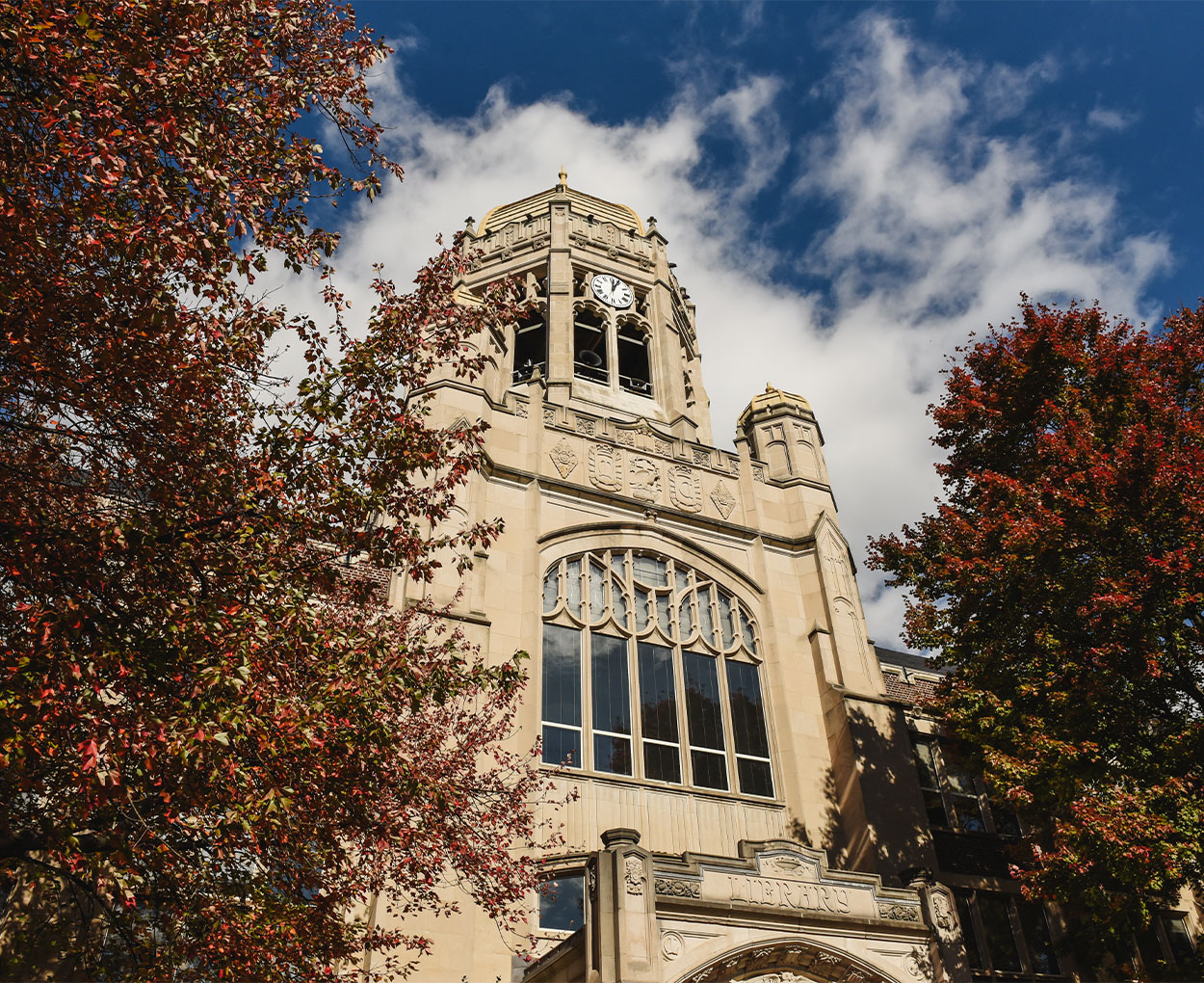 The Haas Clocktower stands against a blue sky dotted with clouds and flanked by trees covered in fall foliage.