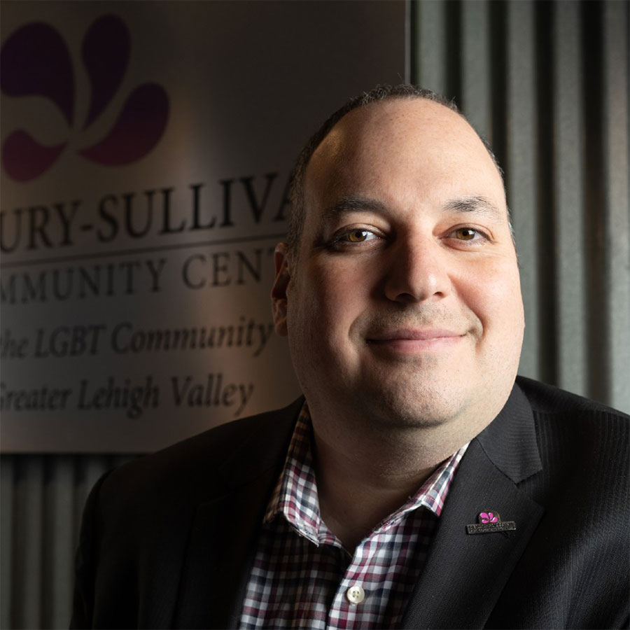 An adult man in a blazer smiles softly at the camera inside a building with a sign that reads 