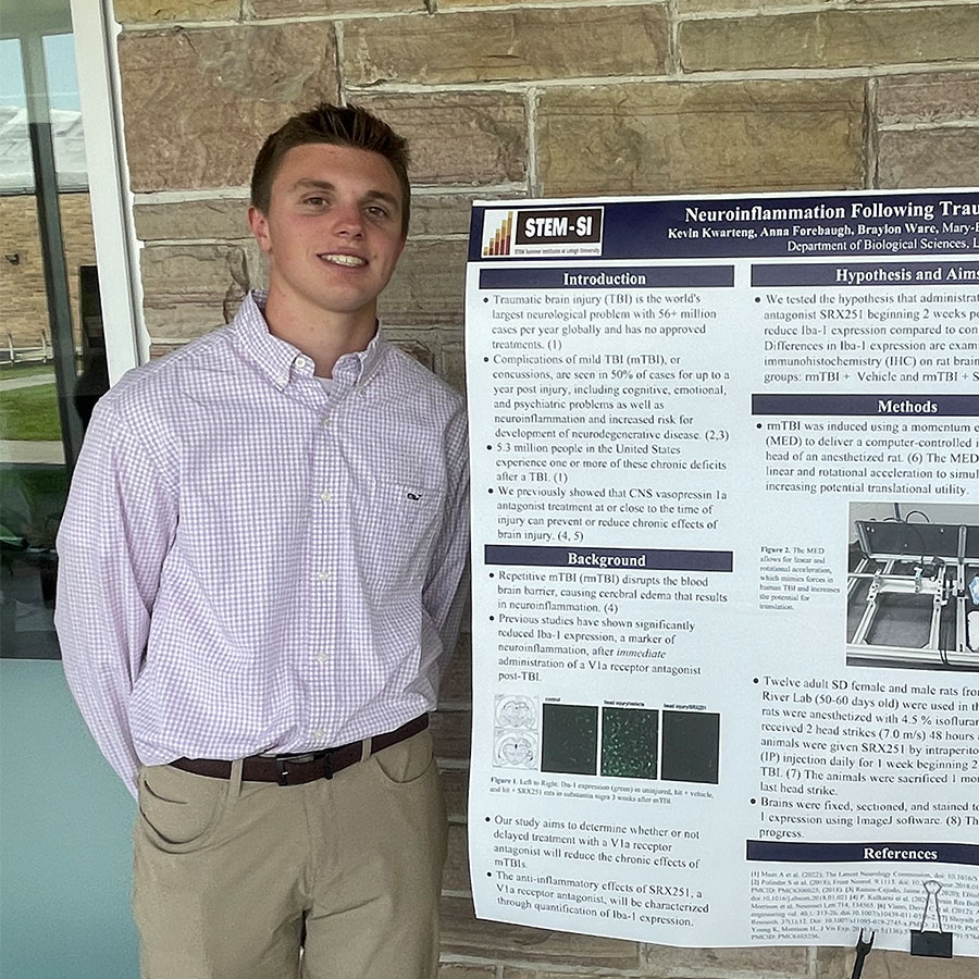 A college student wearing a dress shirt and slacks smiles while standing next to a large poster board with details about a research project.