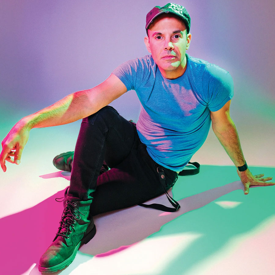 A portrait of a man seated wearing a blue T-shirt and a cap in a room with colorful lighting.