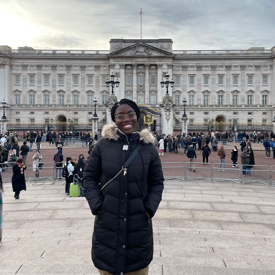 A college student in a black winter coat smiles while standing in front of Buckingham Palace in London.