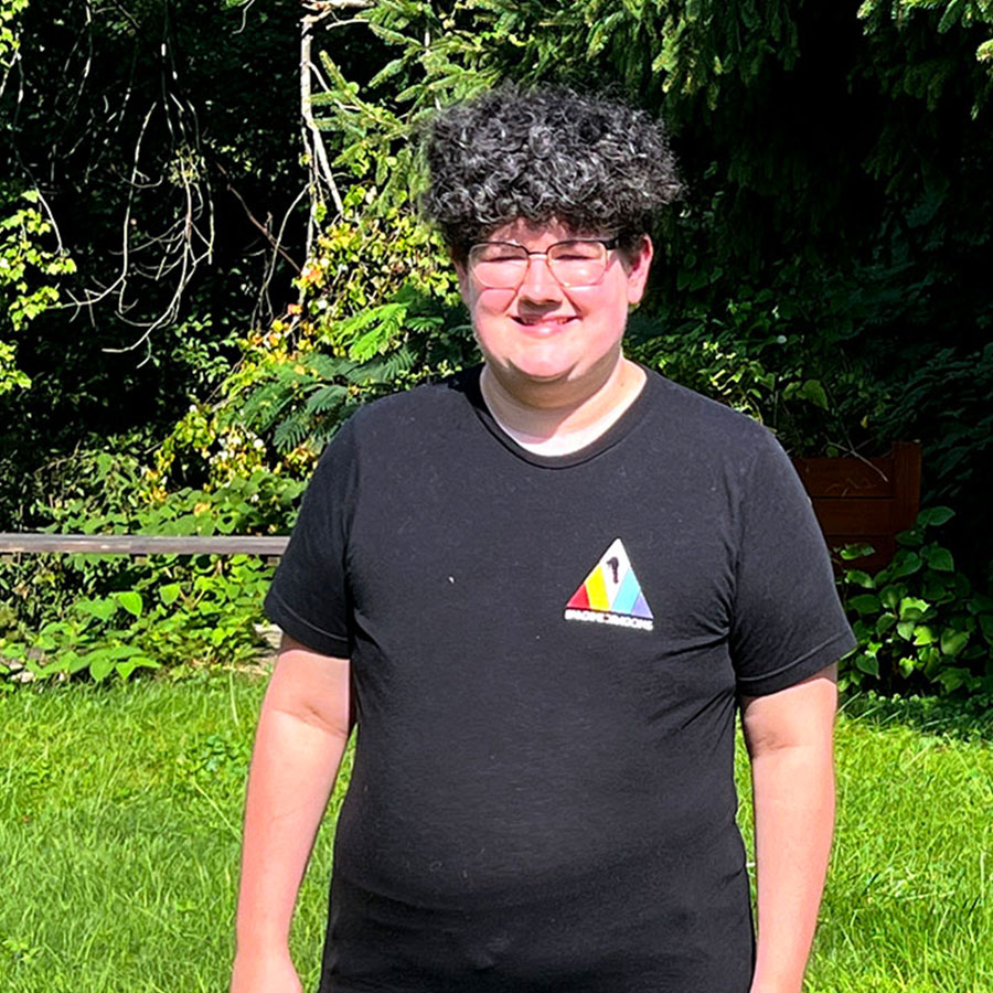 A college student with short, curly hair and glasses grins in the sunlight while outdoors.