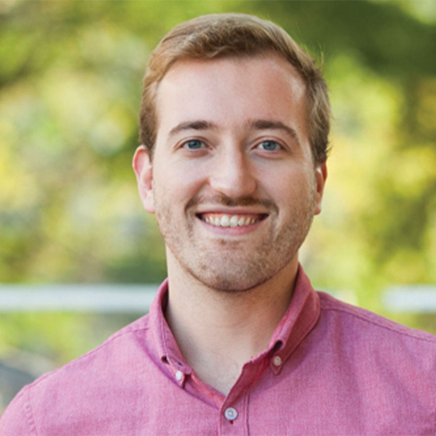 A man in a pink shirt smiles at the camera while outdoors.