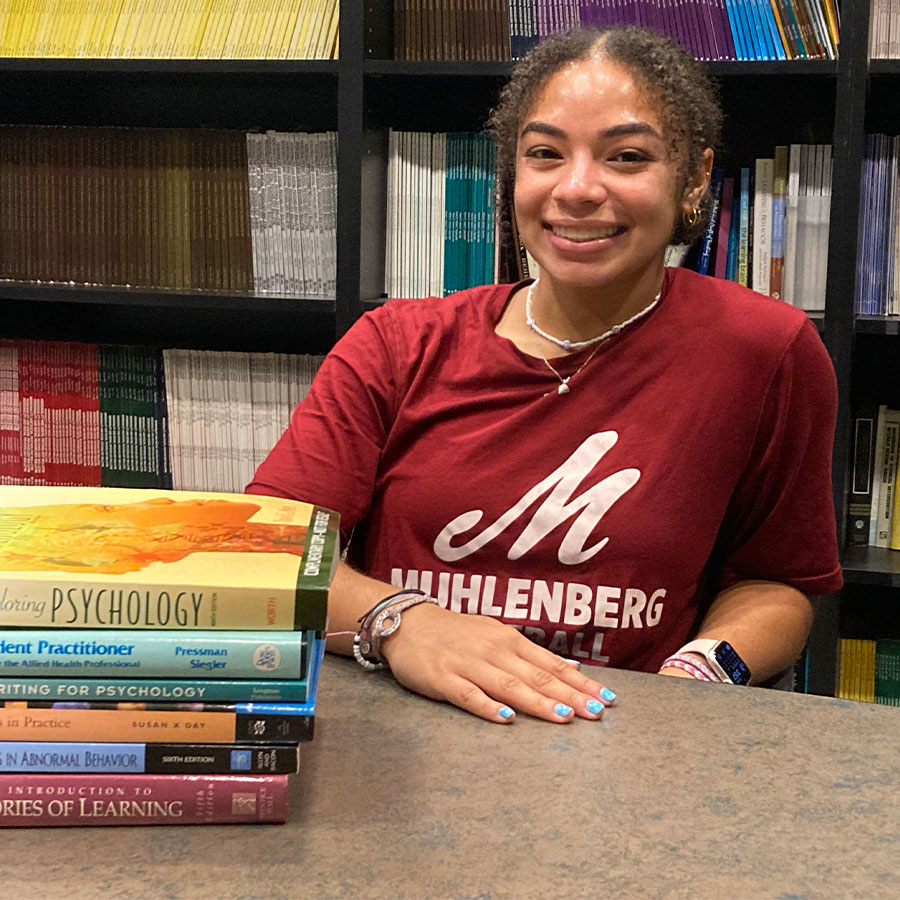 A college students smiles at a desk with a pile of psychology books nearby and a packed bookshelf behind her.