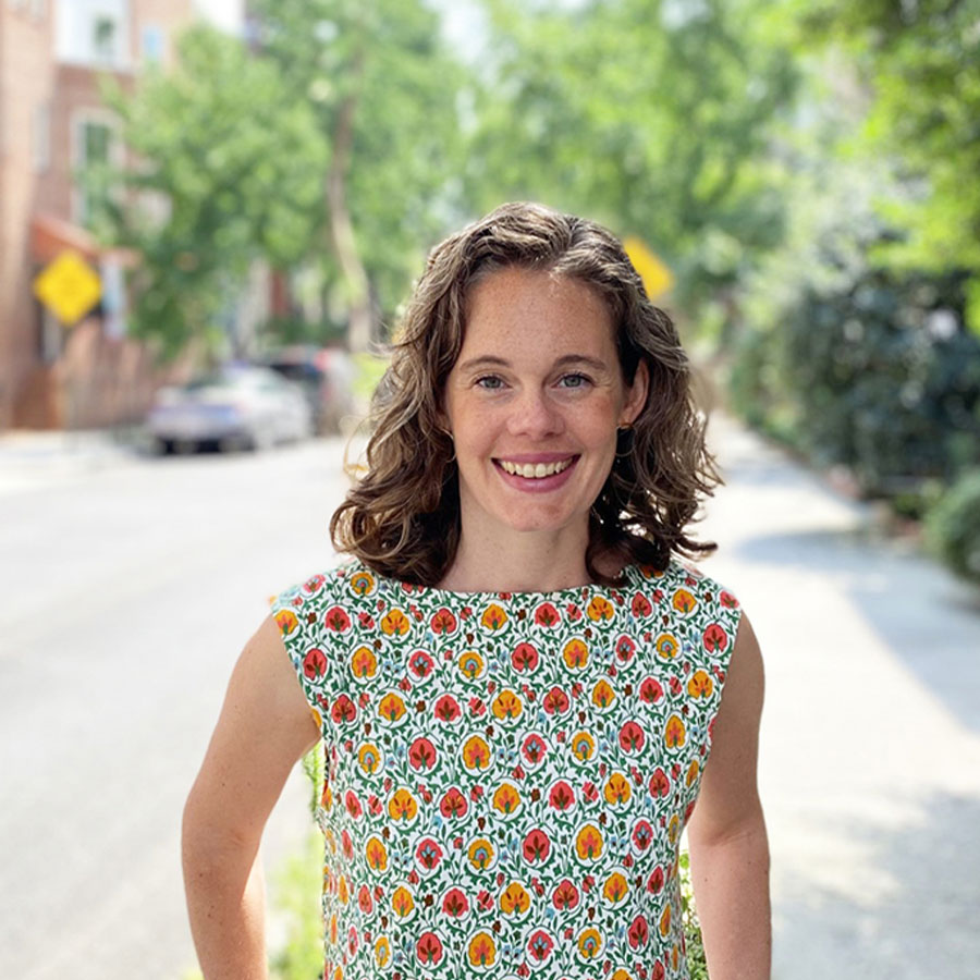 An adult woman in a floral pattern sleeveless blouse stands smiling on a leafy city street.