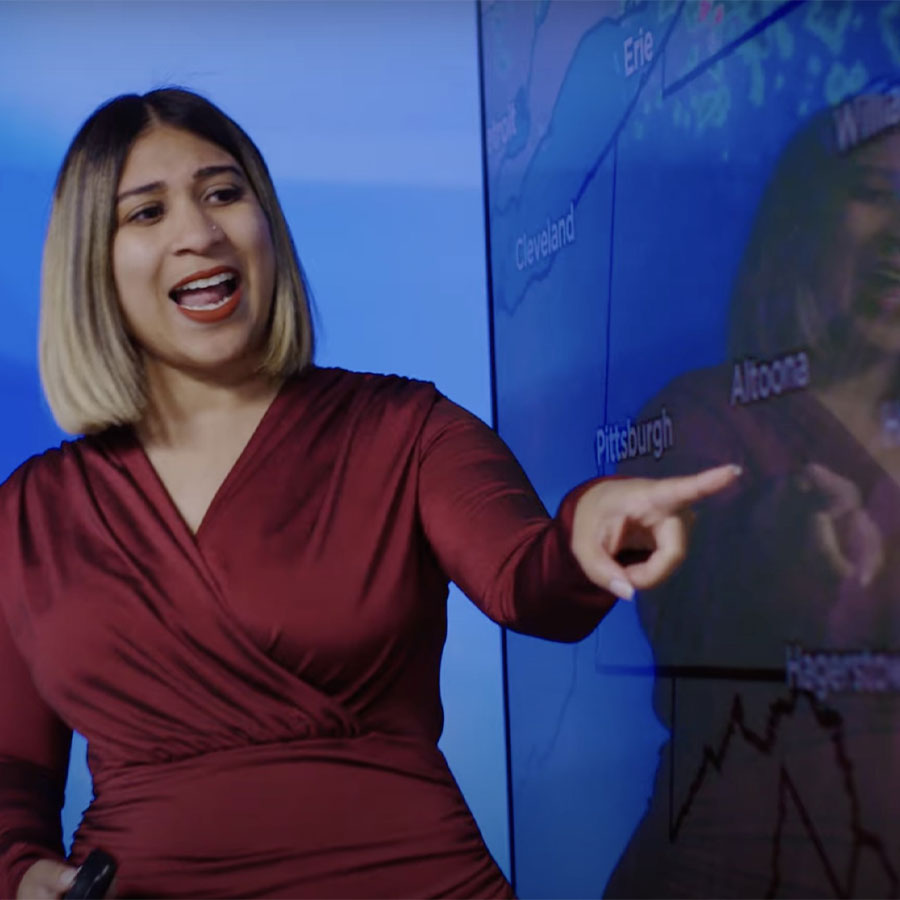 A woman in a red dress speaks while pointing at a digital weather map.