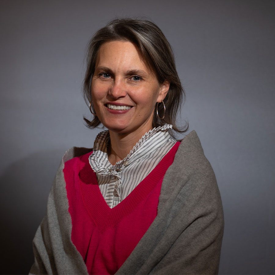 A woman in a red sweater and a grey shawl smiles at the camera in front of a grey backdrop.